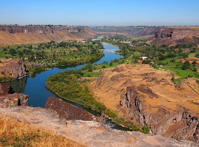 [The river curves into a wide area at the base of the canyon walls. There are many trees and grass in this area as well as several bridges across the water (but not across the canyon).]